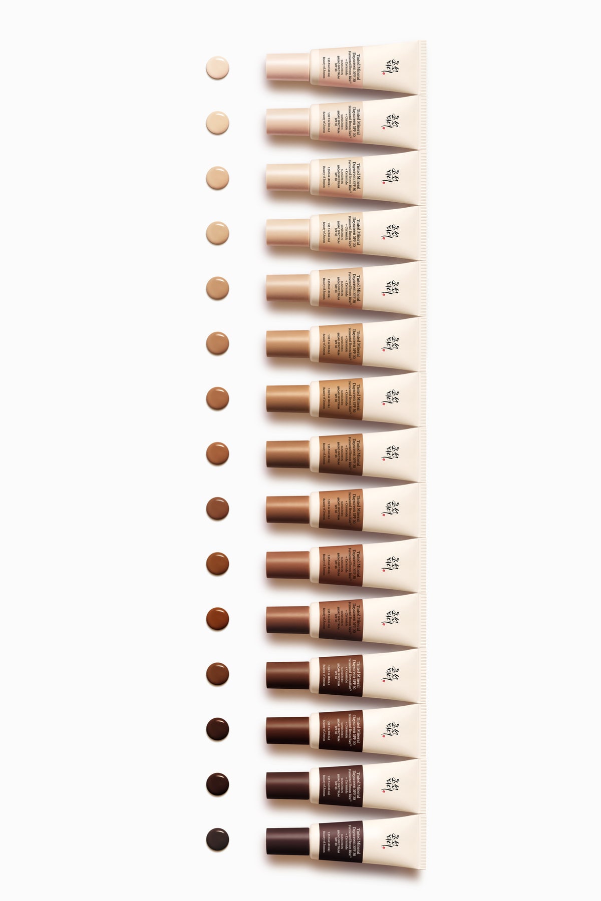 A vertical line of 14 Beauty of Joseon sunscreen tubes lined up from lightest to darkest shade on a white background, each with a matching liquid swatch on the left.